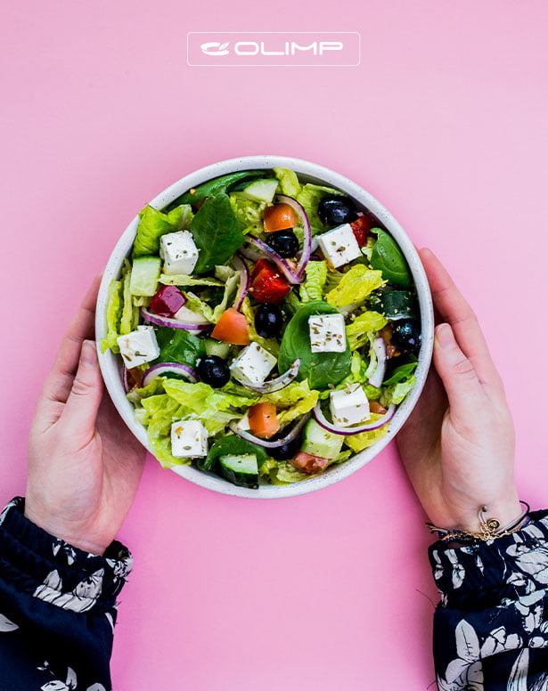 Sałatka grecka z serem feta, oliwkami, pomidorami, ogórkiem i czerwoną cebulą w białej misce, trzymana przez osobę na różowym tle, widoczne logo Olimp na górze.