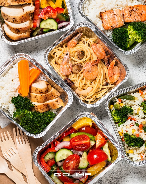 Zestaw różnorodnych dań w aluminiowych pojemnikach: grillowany kurczak, ryba z brokułami, sałatka warzywna i makaron z krewetkami, ułożone na jasnym tle obok drewnianych widelców.