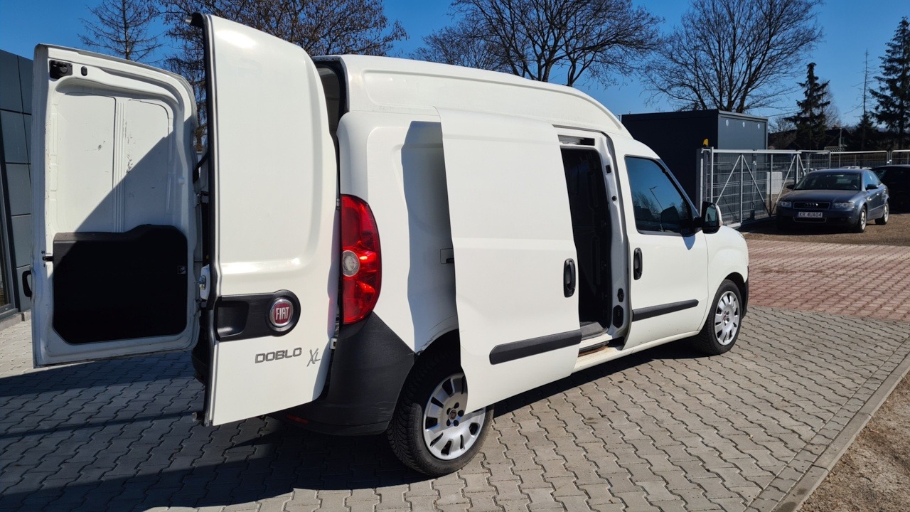 Biały samochód dostawczy Fiat Doblo XL z otwartymi tylnymi i bocznymi drzwiami, zaparkowany na kostce brukowej, w tle budynek i samochód osobowy