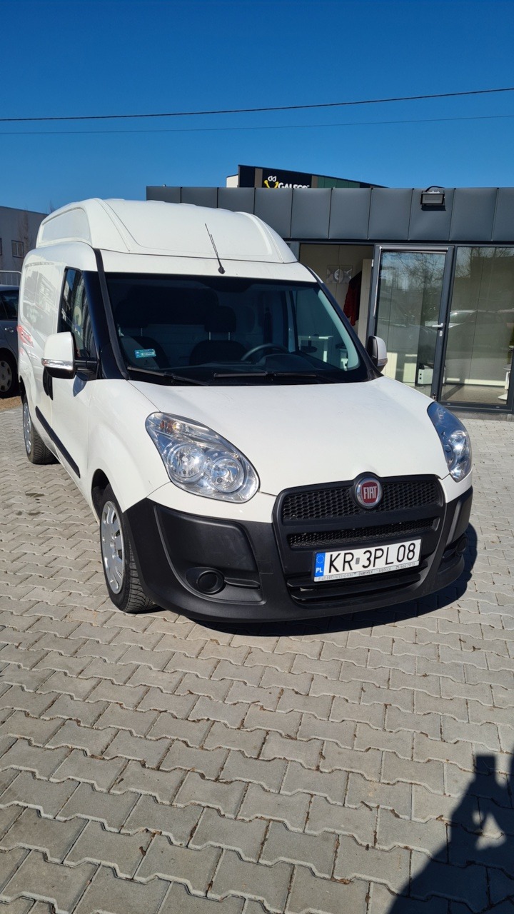 Biały samochód dostawczy Fiat Doblo z wysokim dachem, zaparkowany na brukowanym placu przed budynkiem z logo firmy na dachu, widok z przodu.