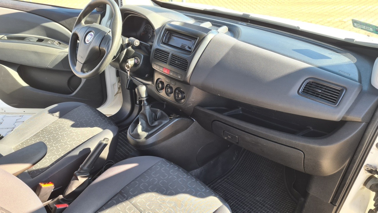 Wnętrze białego samochodu dostawczego Fiat Doblo, widok na deskę rozdzielczą, kierownicę i fotele z materiałową tapicerką w odcieniach szarości.
