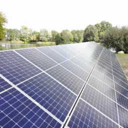 Rozległa instalacja fotowoltaiczna z widokiem na staw i drzewa w tle, panele ustawione pod kątem, odbijające światło.