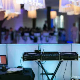 DJ stanowisko z laptopem, mikserem i gramofonami na tle sali weselnej z dekoracjami i oświetleniem w odcieniach błękitu.