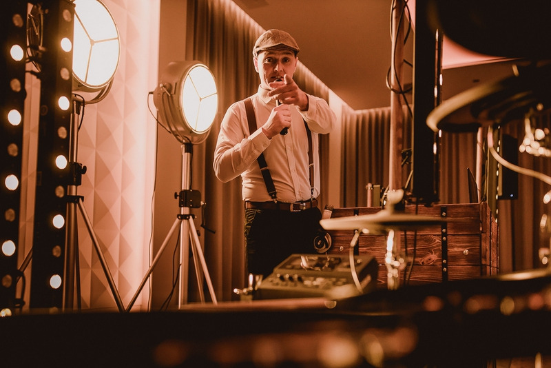 Wokalista w stylu retro z mikrofonem na scenie, oświetlony reflektorami i dekoracjami świetlnymi, częściowo widoczny zestaw perkusyjny.