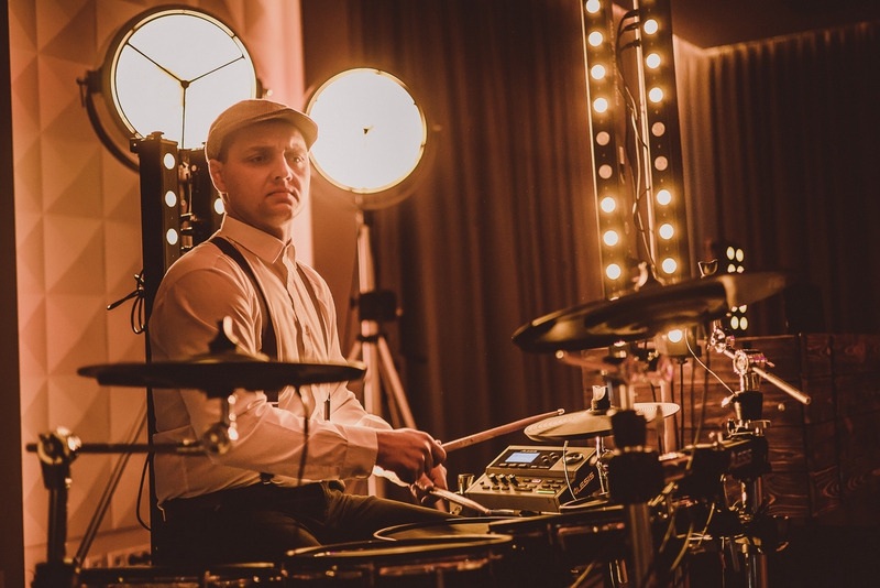 Perkusista w kaszkiecie i szelkach gra na perkusji elektronicznej Alesis, oświetlony przez reflektory sceniczne w ciepłych barwach.