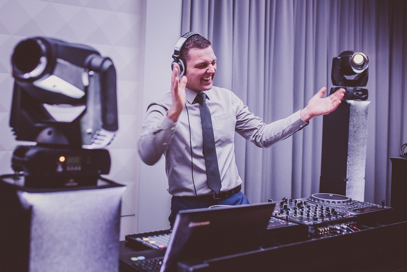 Uśmiechnięty DJ w słuchawkach, w jasnej koszuli i krawacie, gestykuluje rękami przed mikserem DJ-skim i oświetleniem scenicznym.