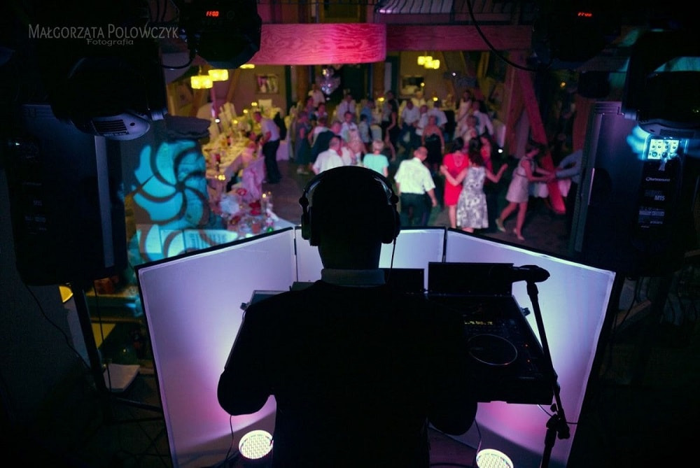 Sylwetka DJ-a w słuchawkach za konsoletą podczas imprezy tanecznej, w tle bawiący się goście i dekoracyjne oświetlenie.