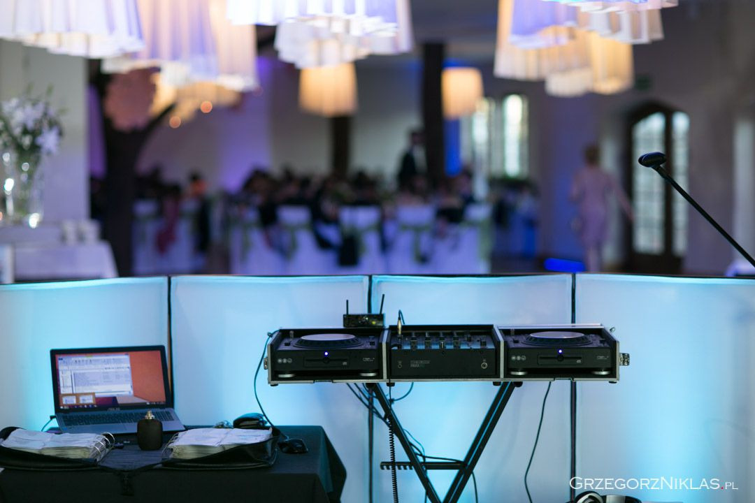 DJ stanowisko z laptopem, mikserem i gramofonami na tle sali weselnej z dekoracjami i oświetleniem w odcieniach błękitu.