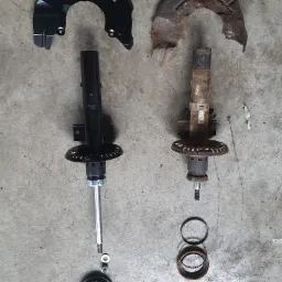 Porównanie nowego i zużytego amortyzatora samochodowego z osłoną tarczy hamulcowej, widoczne ślady korozji na starym elemencie.