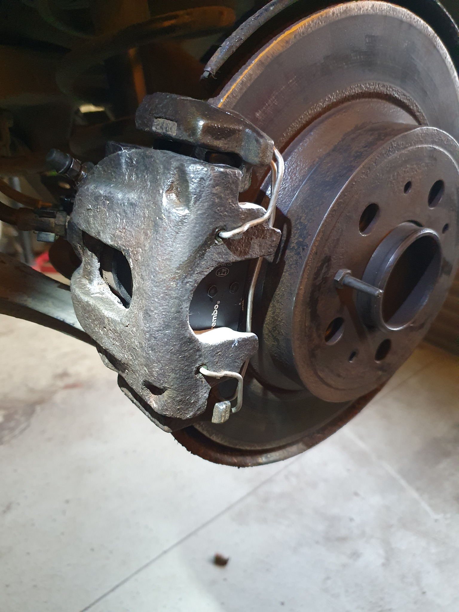 Zużyty zacisk hamulcowy z nowymi klockami marki Brembo, widoczna tarcza hamulcowa z otworami i śrubą mocującą, ujęcie z bliska.