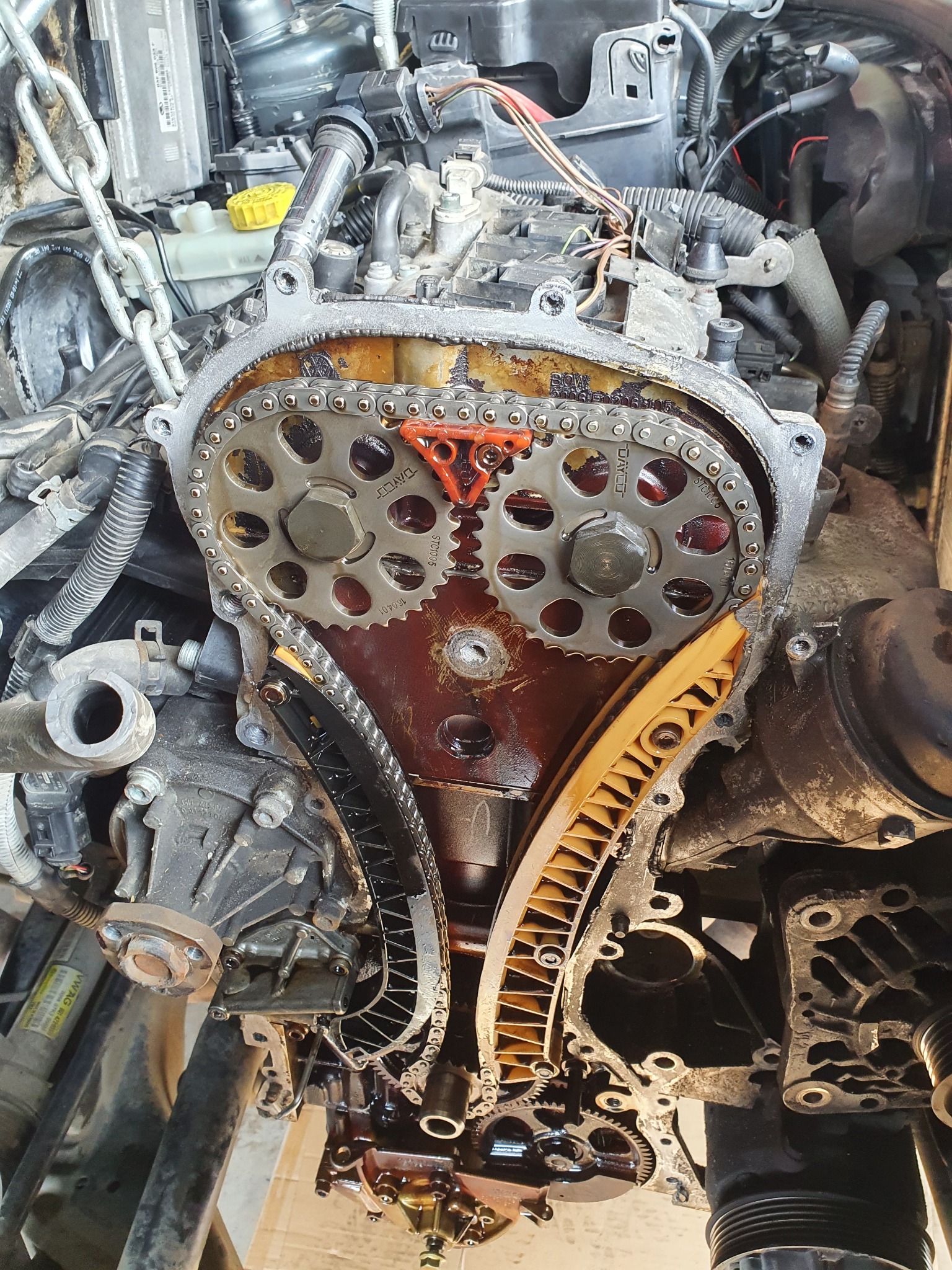 Otwarty silnik samochodowy z widocznym łańcuchem rozrządu, zębatkami i prowadnicami, z wyraźnymi śladami oleju i zużycia, ukazujący proces naprawy lub diagnostyki.