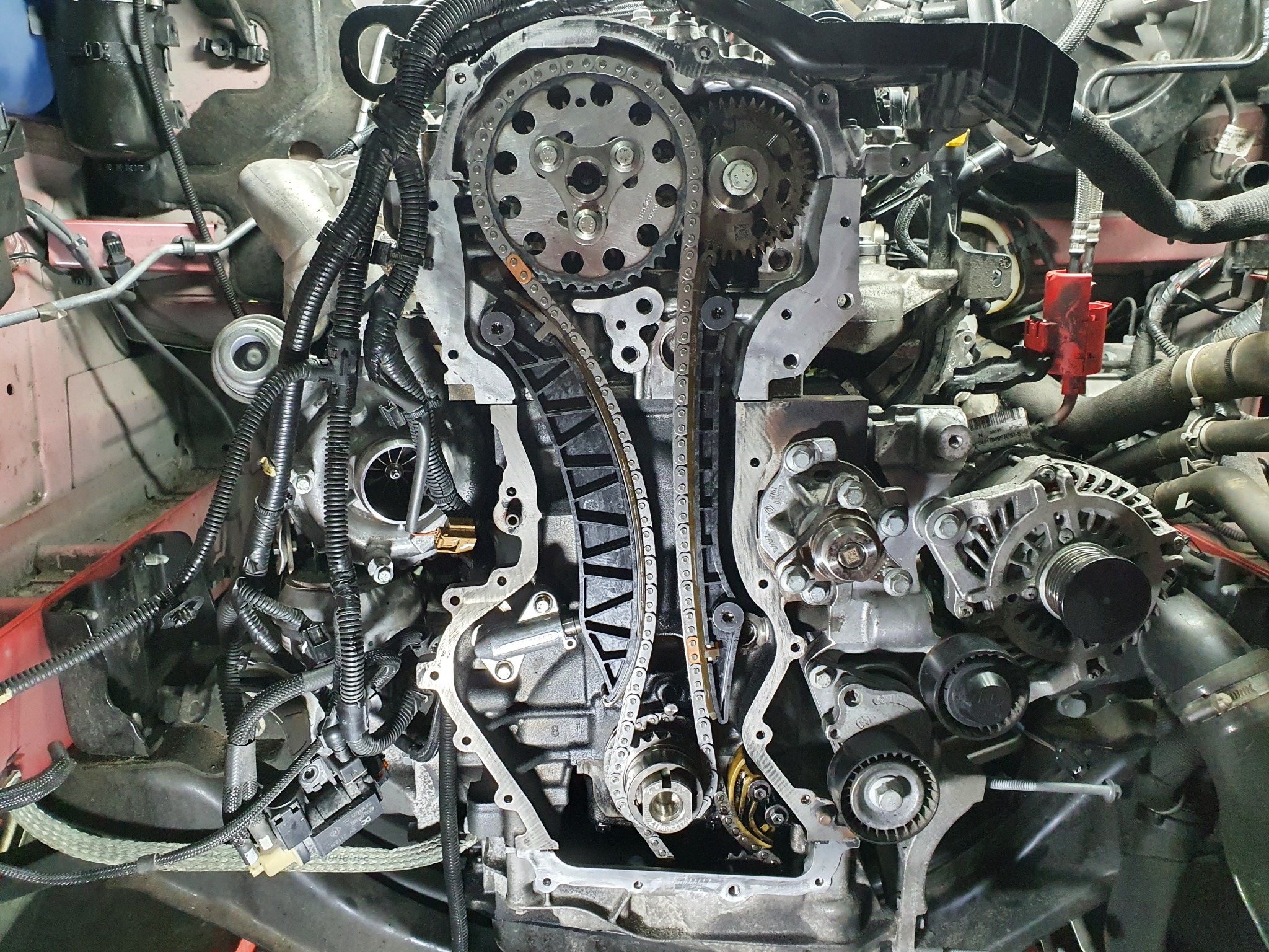 Odsłonięty silnik samochodowy z widocznym łańcuchem rozrządu, kołami zębatymi i wiązkami przewodów, ukazujący skomplikowaną konstrukcję wewnętrzną.