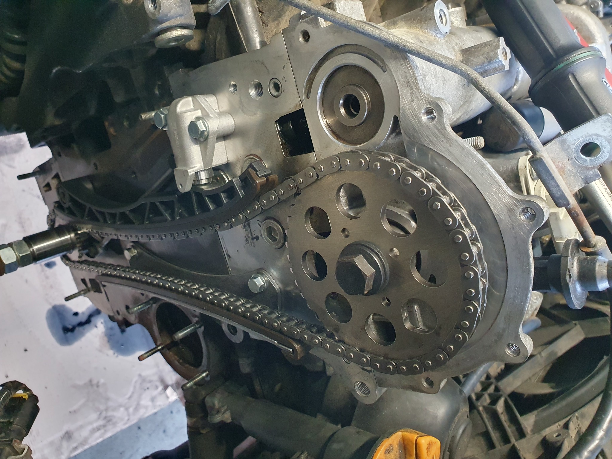 Widok z bliska na rozmontowany silnik samochodowy z łańcuchem rozrządu i kołem zębatym, widoczne ślady oleju, nakrętki i śruby montażowe.