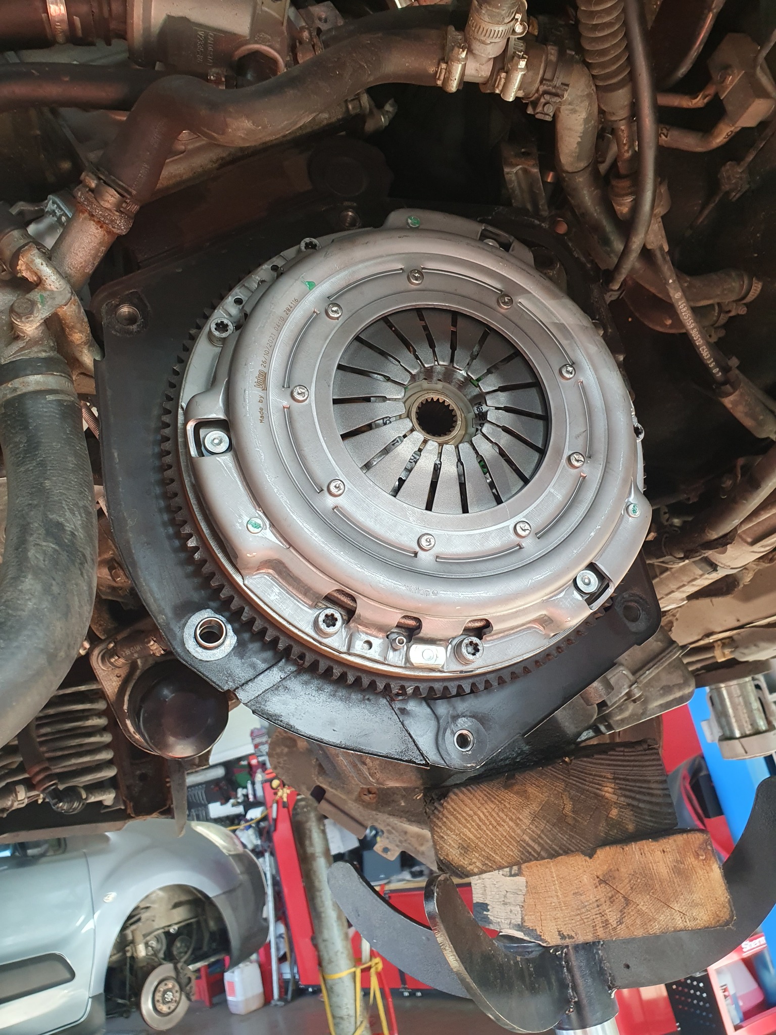 Widok od dołu na sprzęgło samochodowe zamontowane w pojeździe, podparte drewnianymi klockami na podnośniku, widoczne elementy silnika i zawieszenia, w tle fragment innego samochodu bez koła.