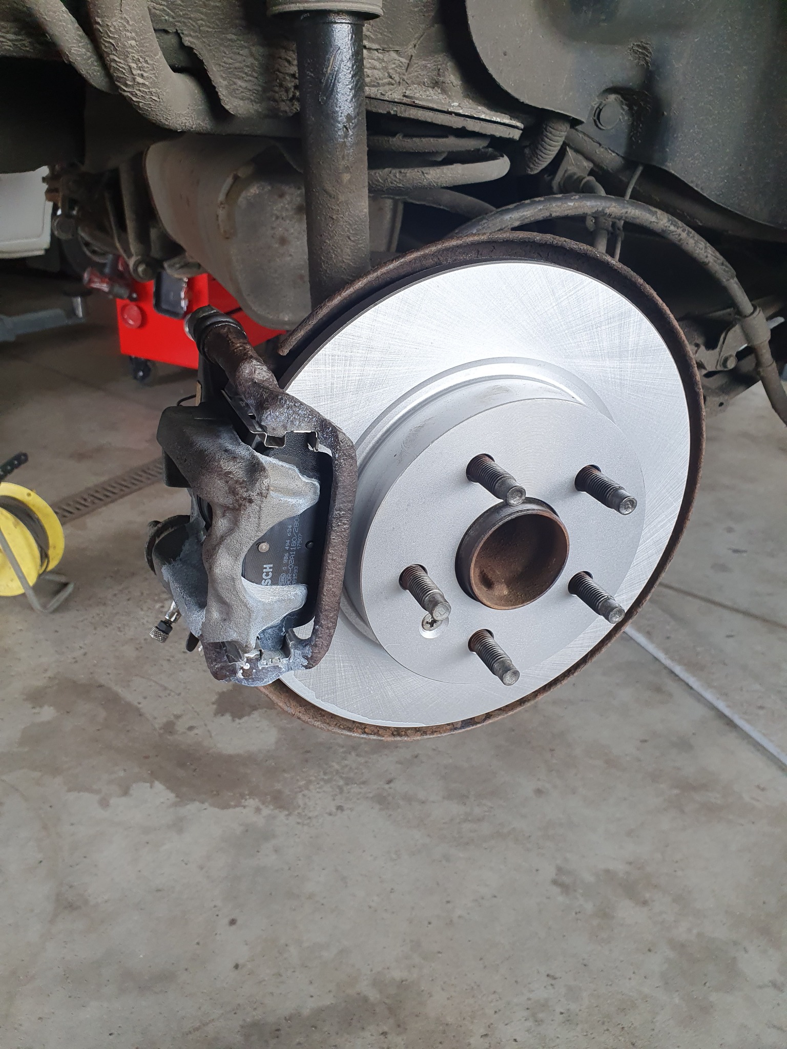 Nowa tarcza hamulcowa zamontowana na piaście, widoczny zacisk hamulcowy z klockami marki Bosch, tło stanowią elementy zawieszenia pojazdu i podnośnik warsztatowy.