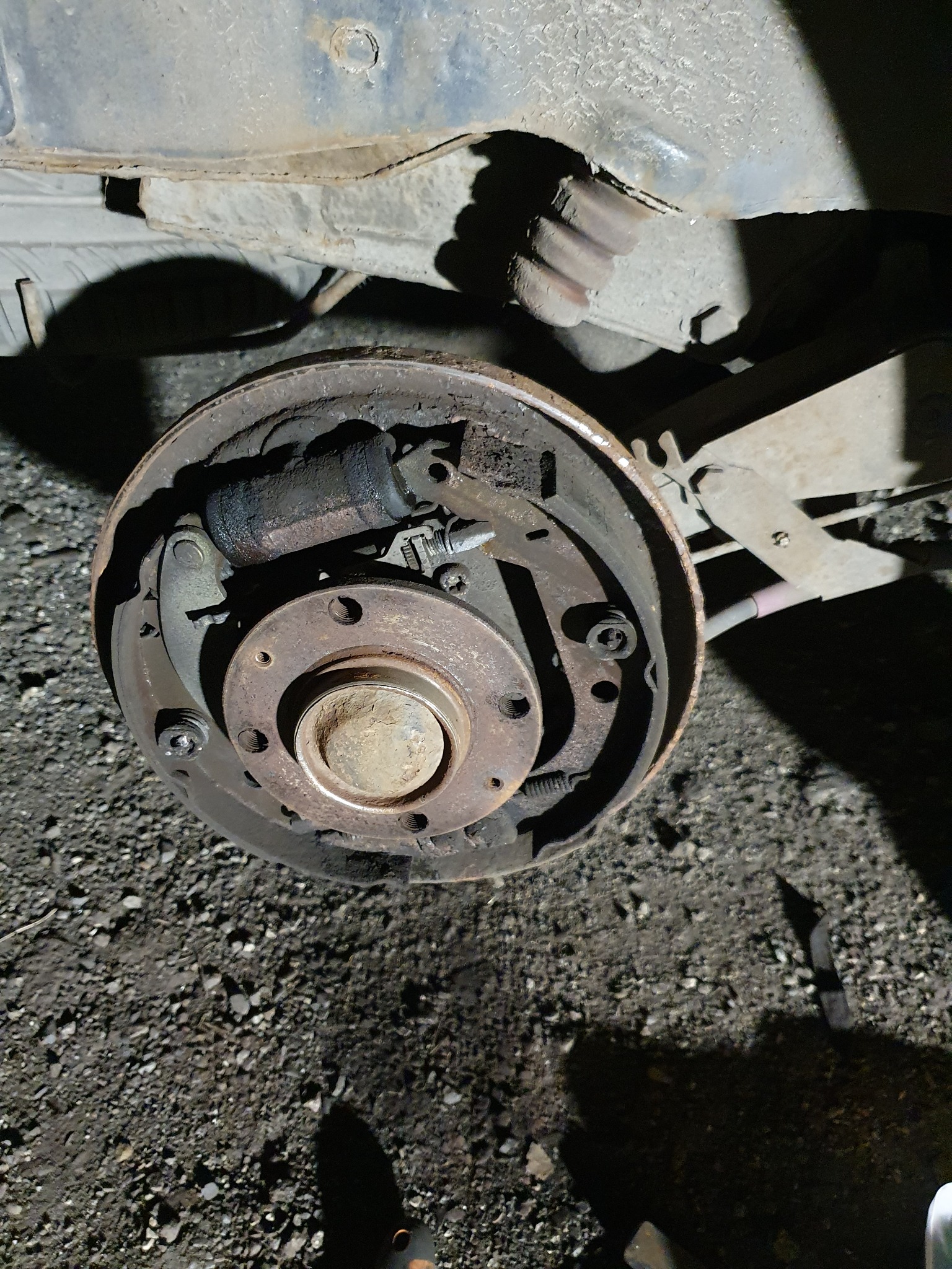 Otwarty bęben hamulcowy z widocznymi szczękami, cylinderkiem i sprężynami, sfotografowany z bliska na tle podłoża żwirowego.