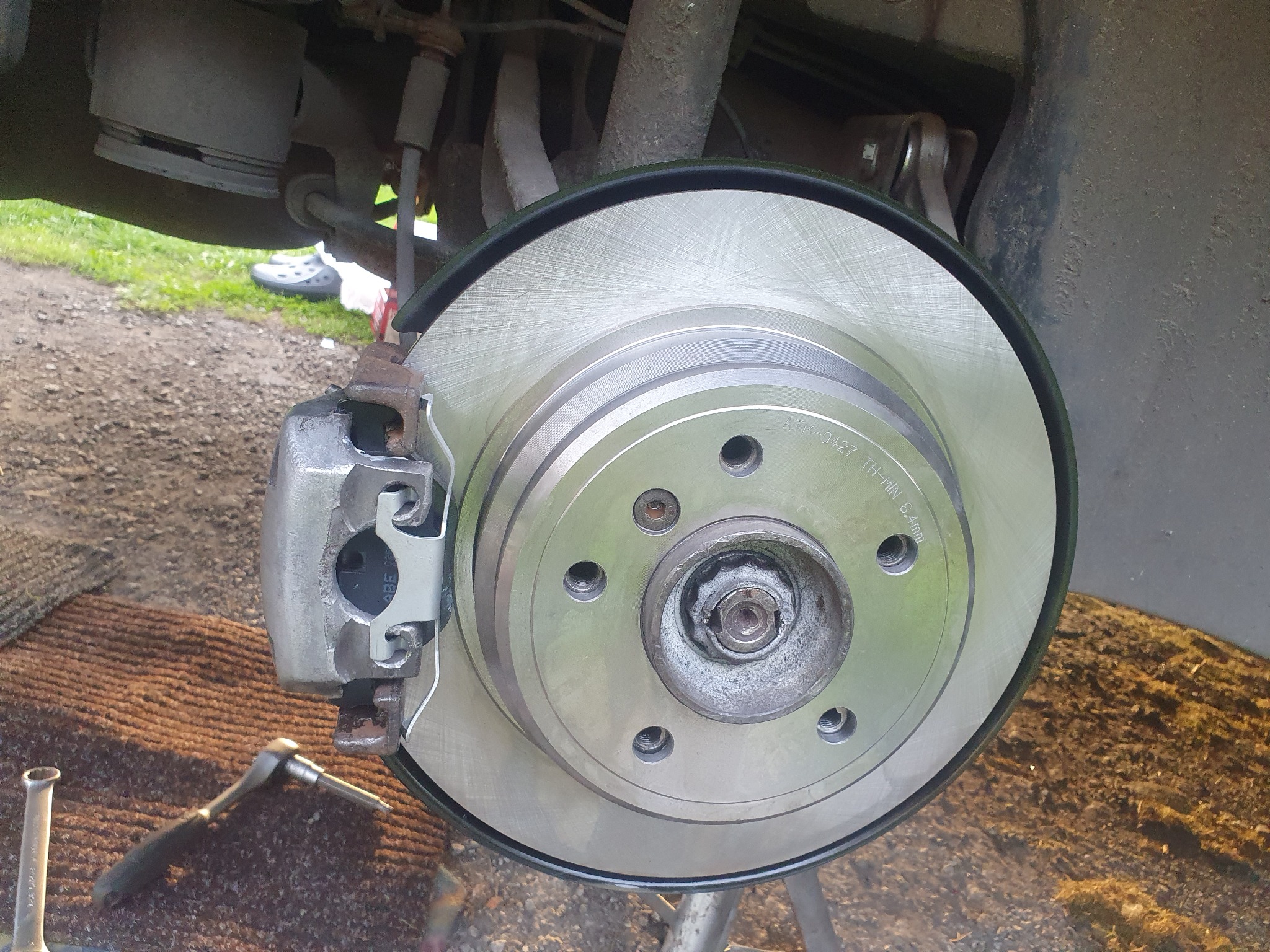 Nowa tarcza hamulcowa z zaciskiem, widoczne narzędzia i fragment podwozia pojazdu.