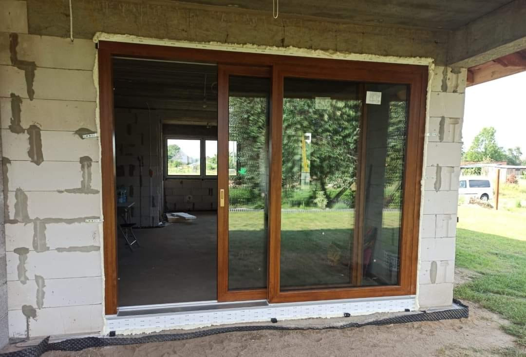 Brązowe przesuwne okno tarasowe w surowym stanie, widoczne bloczki betonowe i elementy wykończeniowe, trawa na zewnątrz.