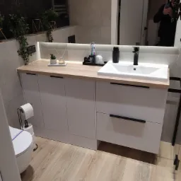 Nowoczesna łazienka z białymi szafkami pod umywalką z drewnianym blatem, czarną armaturą i dekoracjami roślinnymi, widoczne heksagonalne płytki na ścianie i odbicie osoby robiącej zdjęcie w lustrze.