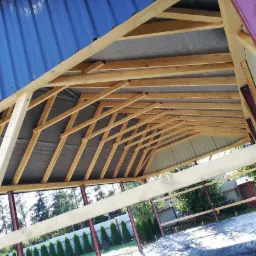 Wnętrze konstrukcji dachu z drewnianą więźbą, widoczne spod spodu, z zamontowaną membraną dachową i fragmentami blachy trapezowej w kolorze niebieskim oraz metalowe słupy nośne pomalowane na bordowo.