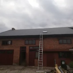 Ciemnoszary dach z blachy falistej na ceglanym budynku z drabiną opartą o dach, widoczne okno dachowe i komin.