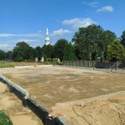 DANPOL USŁUGI KOPARKO-ŁADOWARKĄ, TRANSPORT - Przygotowany teren pod budowę z widocznymi fundamentami, zabezpieczeniem przeciwwilgociowym i częściowo usuniętą ziemią, w tle kościół i zabudowa jednorodzinna.