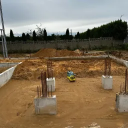 Fundamenty domu jednorodzinnego w trakcie budowy, widoczna ubijarka wibracyjna do zagęszczania podłoża, wystające pręty zbrojeniowe, betonowe ściany fundamentowe i hałda ziemi w tle.