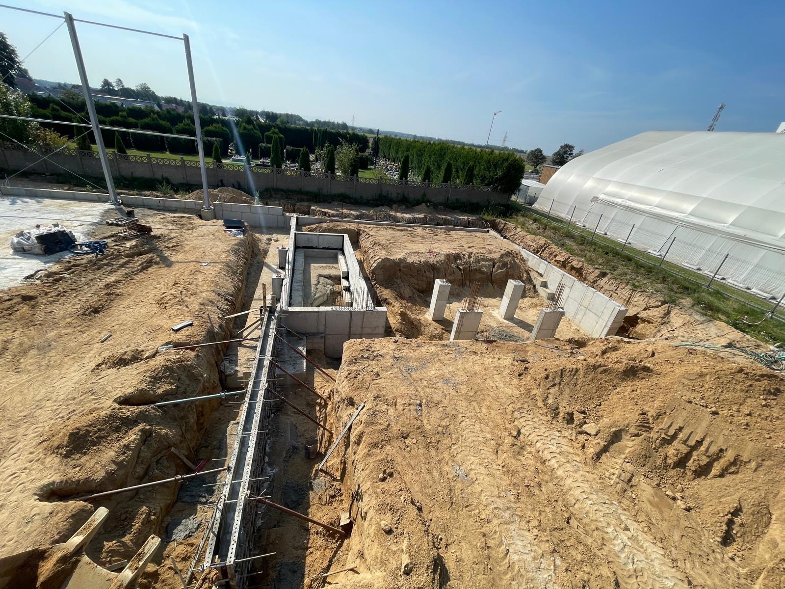 Wykopane fundamenty pod budynek z widocznymi zbrojeniami, szalunkami i słupami betonowymi na tle ziemi i zieleni. W tle widoczny biały namiot magazynowy.