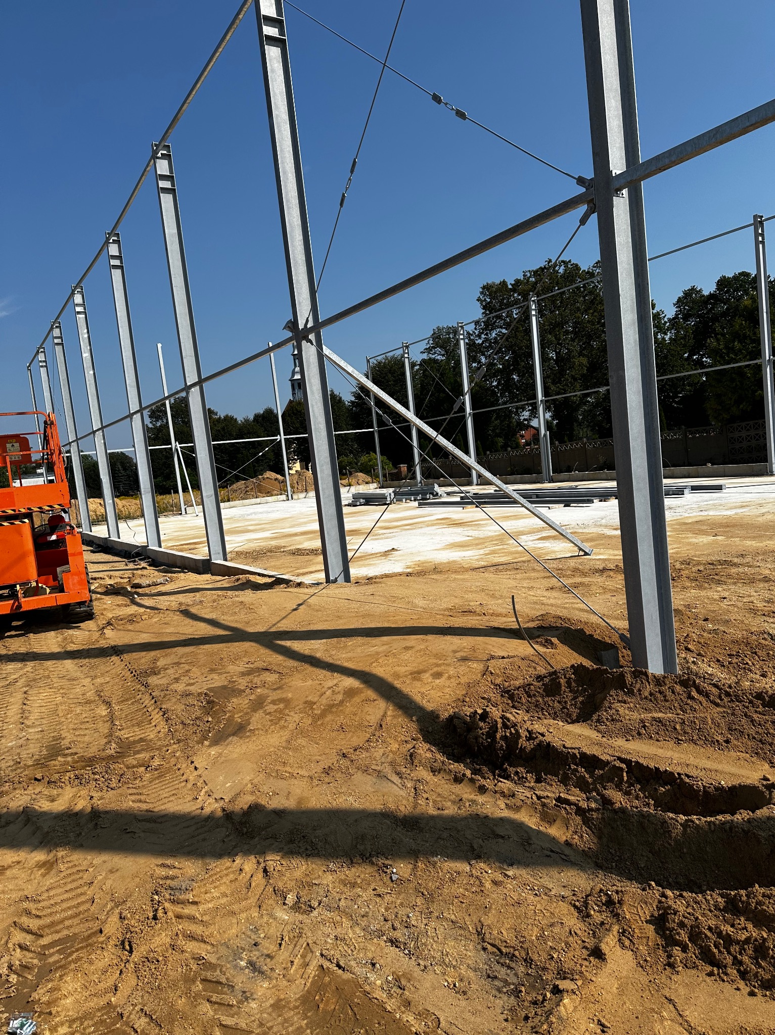 Montaż stalowej konstrukcji hali na wyrównanym terenie, widoczny podnośnik koszowy i fragment fundamentów, w tle drzewa i kościół.