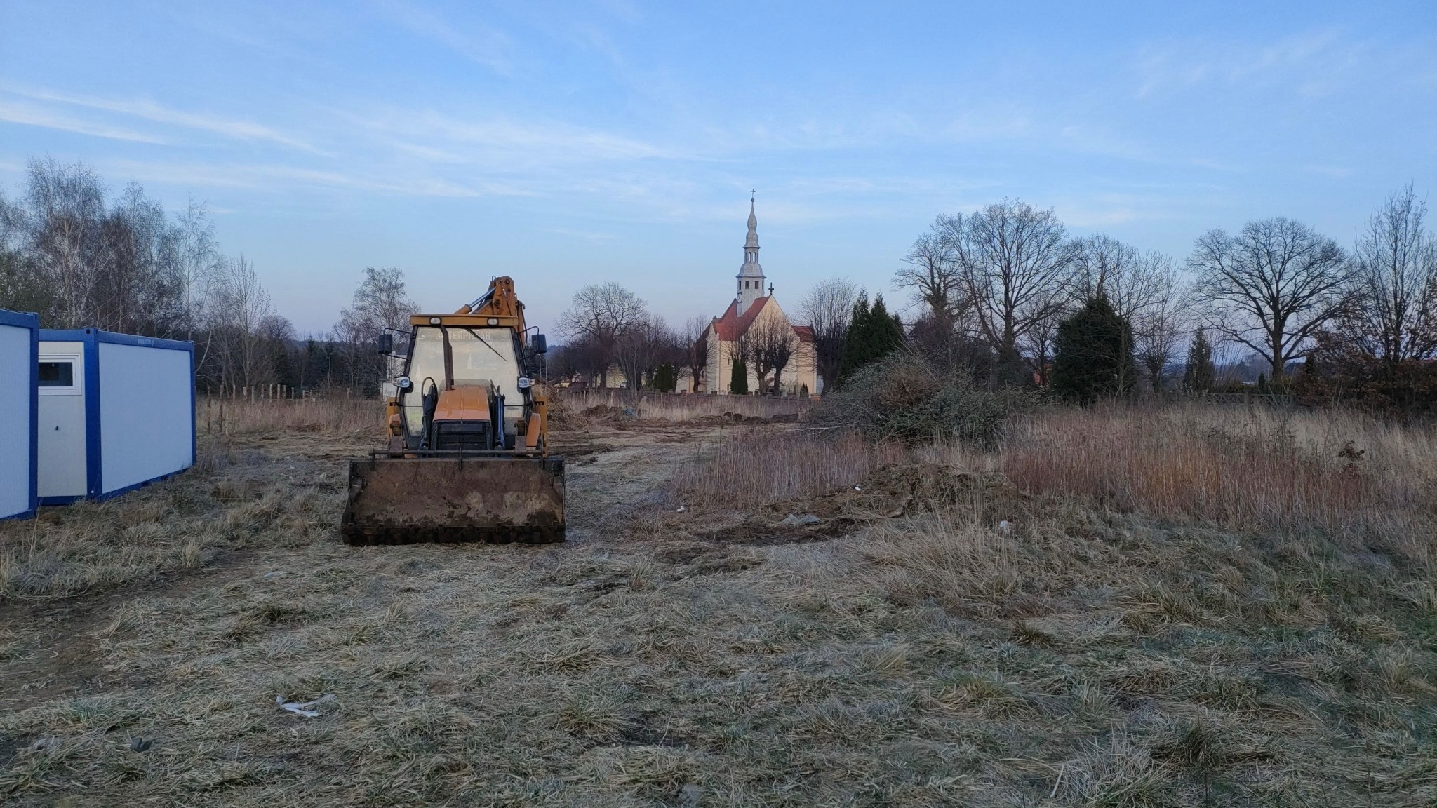 Żółta koparko-ładowarka na działce z wysoką trawą, w tle widoczny kościół i kontenery budowlane, poranna sceneria.
