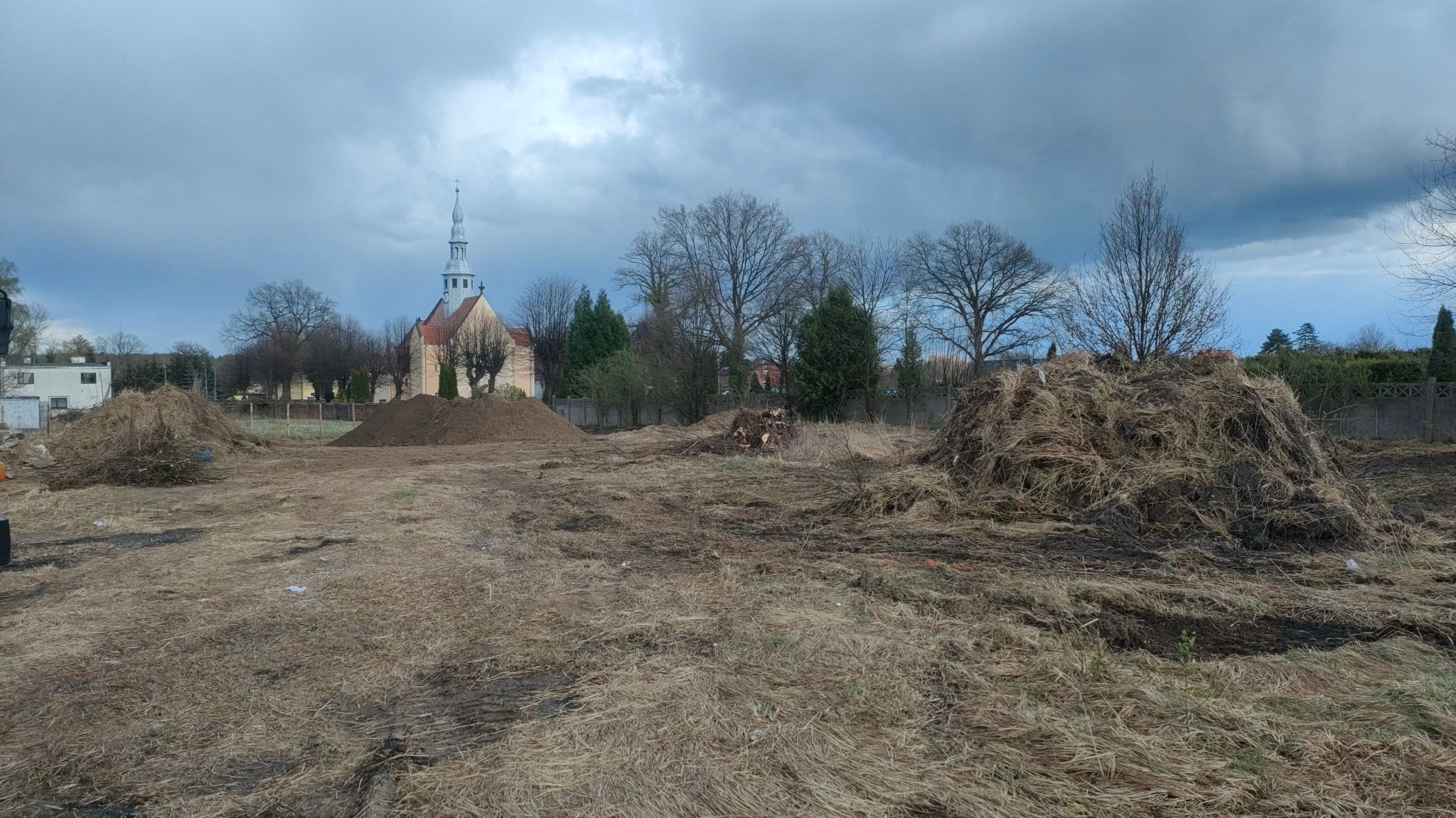 Przygotowany teren pod budowę z widocznymi stertami ziemi i suchych traw, w tle kościół z wysoką wieżą oraz pochmurne niebo.