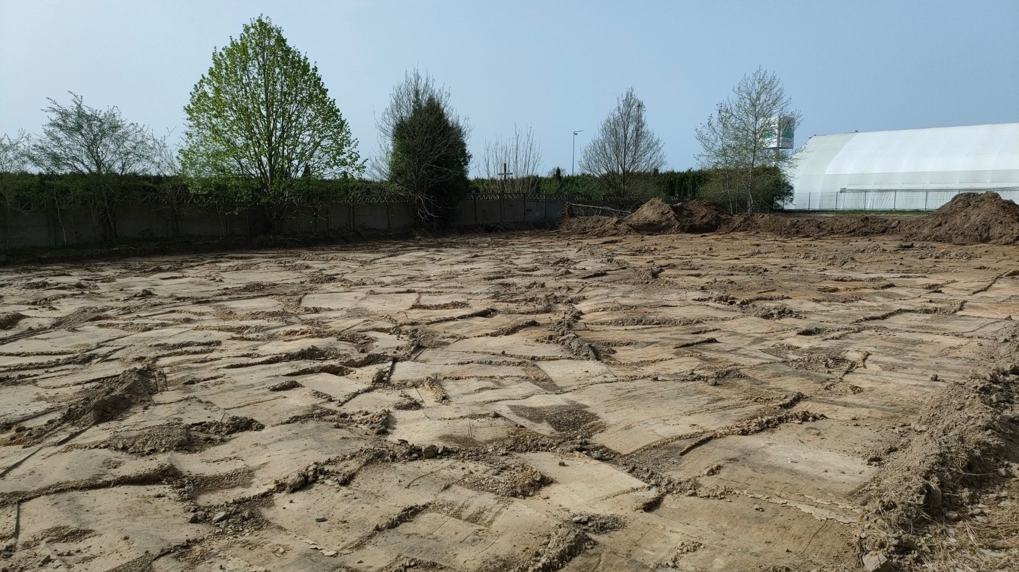 Przygotowany teren pod budowę z widocznymi śladami po maszynach, w tle zieleń i biały namiot.