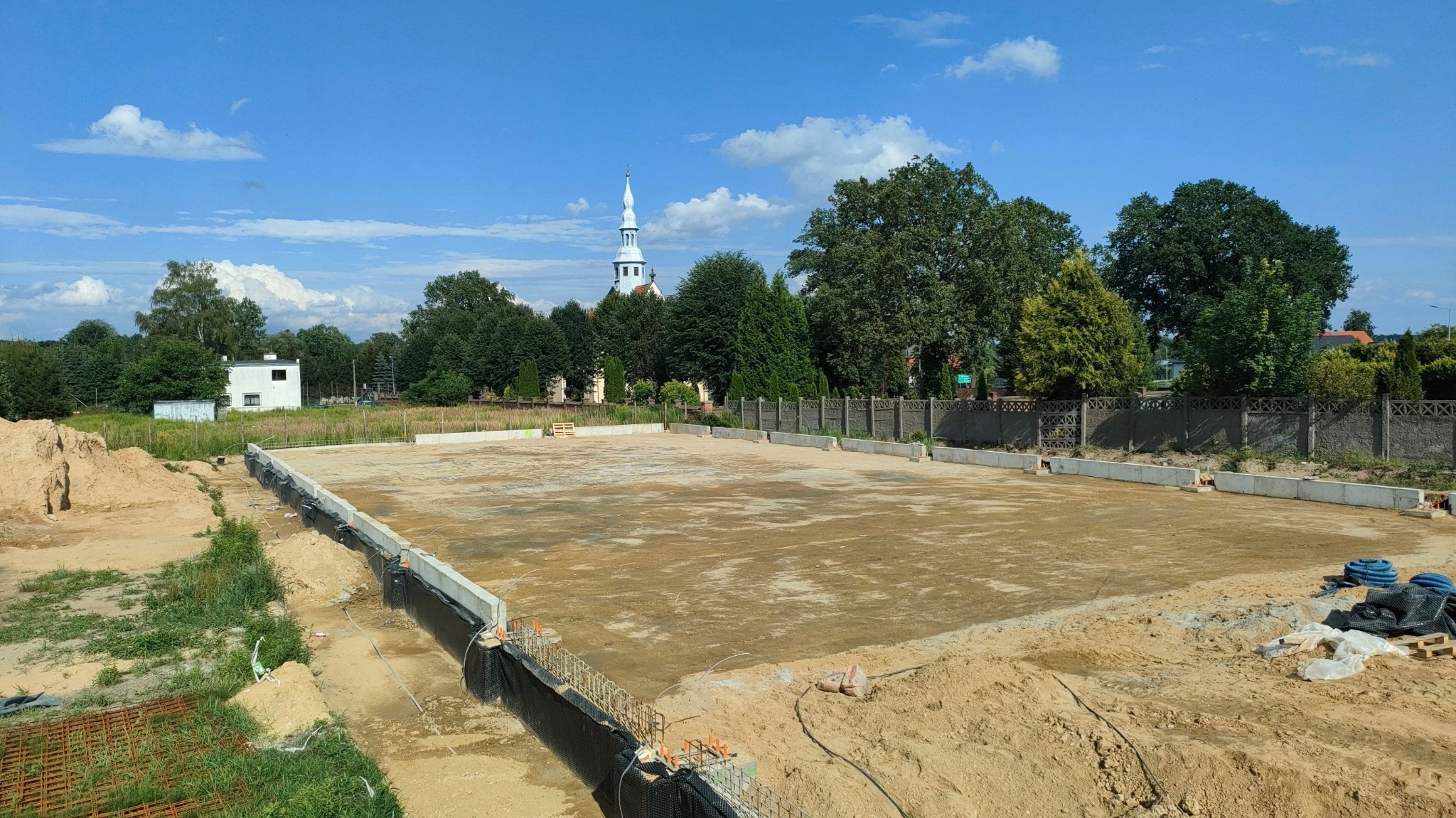 Przygotowany teren pod budowę z widocznymi fundamentami, zabezpieczeniem przeciwwilgociowym i częściowo usuniętą ziemią, w tle kościół i zabudowa jednorodzinna.