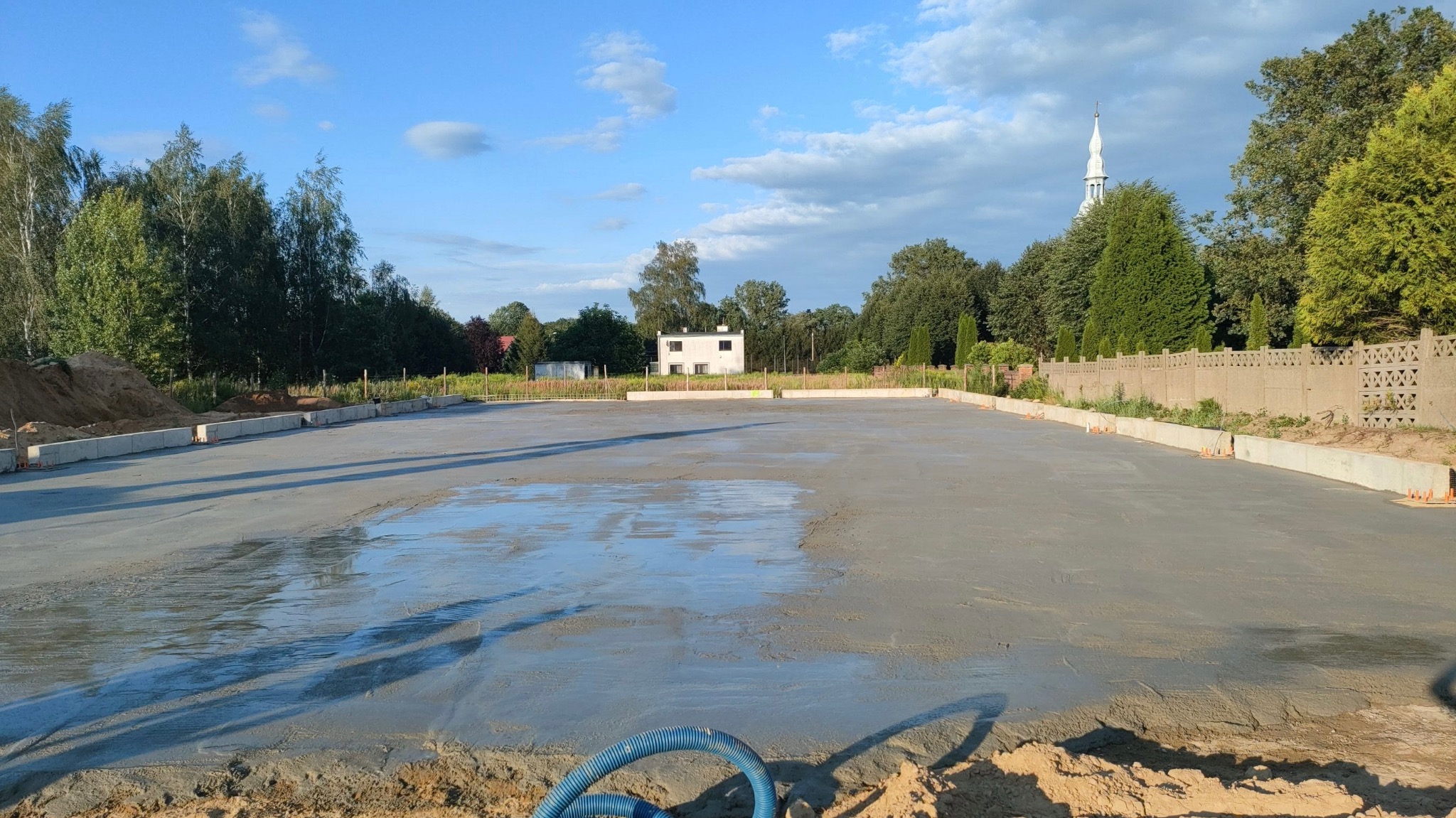 Przygotowany, mokry betonowy podkład pod budowę z widocznymi betonowymi krawężnikami, w tle budynek i drzewa.