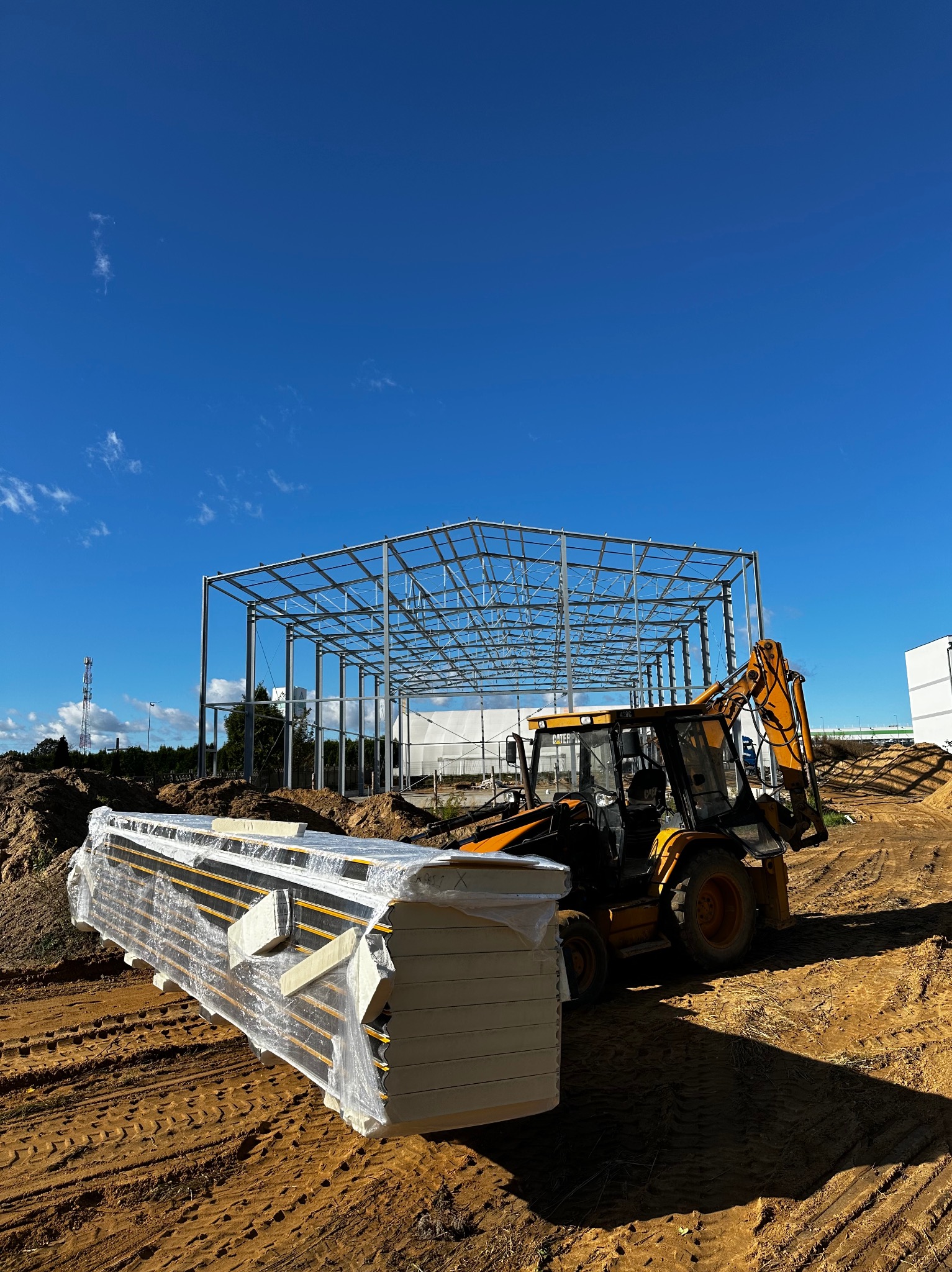 Żółta koparko-ładowarka transportuje owinięte folią płyty warstwowe na placu budowy z widoczną stalową konstrukcją budynku na tle błękitnego nieba.