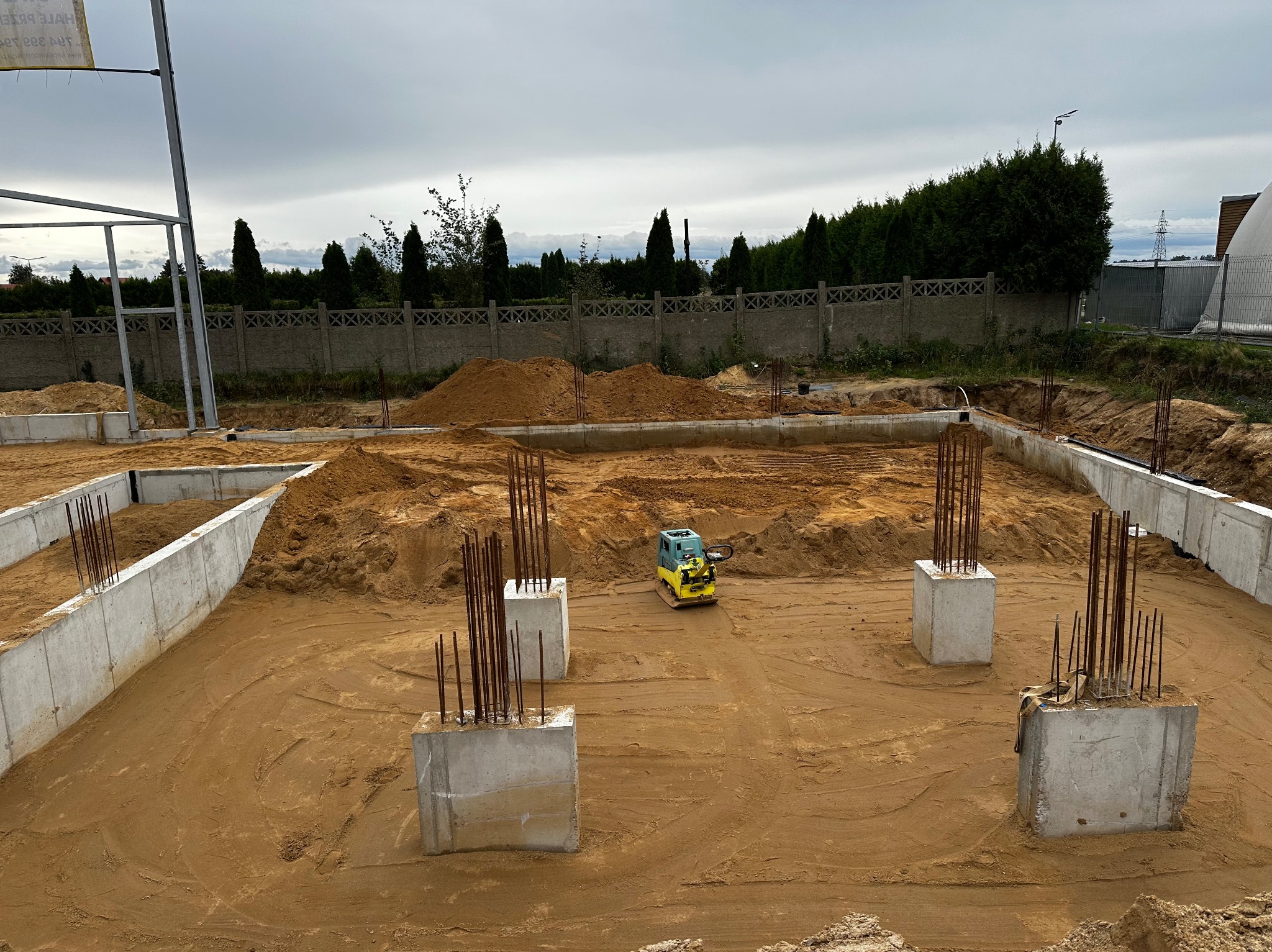 Fundamenty domu jednorodzinnego w trakcie budowy, widoczna ubijarka wibracyjna do zagęszczania podłoża, wystające pręty zbrojeniowe, betonowe ściany fundamentowe i hałda ziemi w tle.