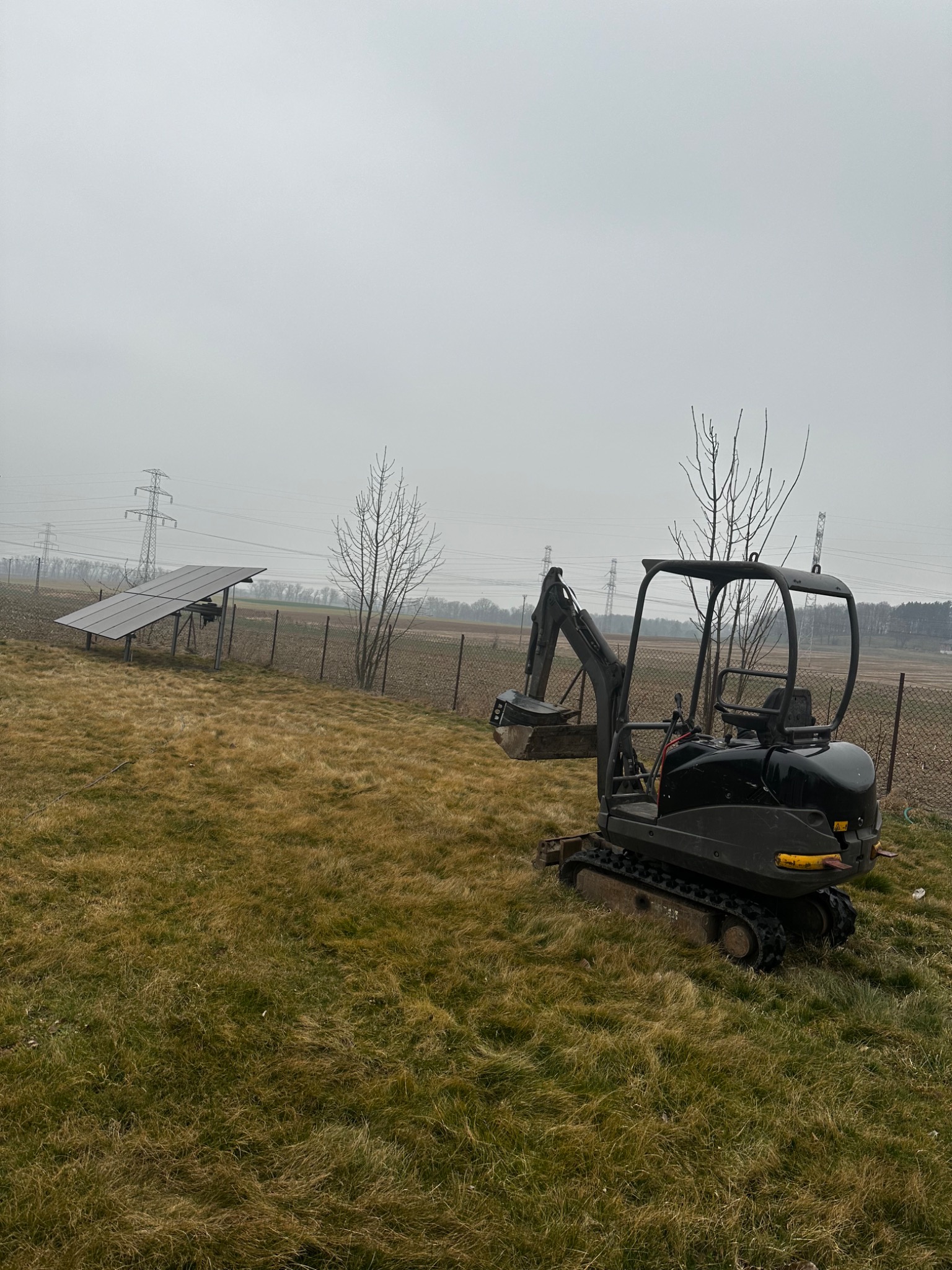 Mała czarna koparka gąsienicowa zaparkowana na zewnątrz na trawiastym terenie, z panelami słonecznymi i liniami energetycznymi w tle, w pochmurny dzień.