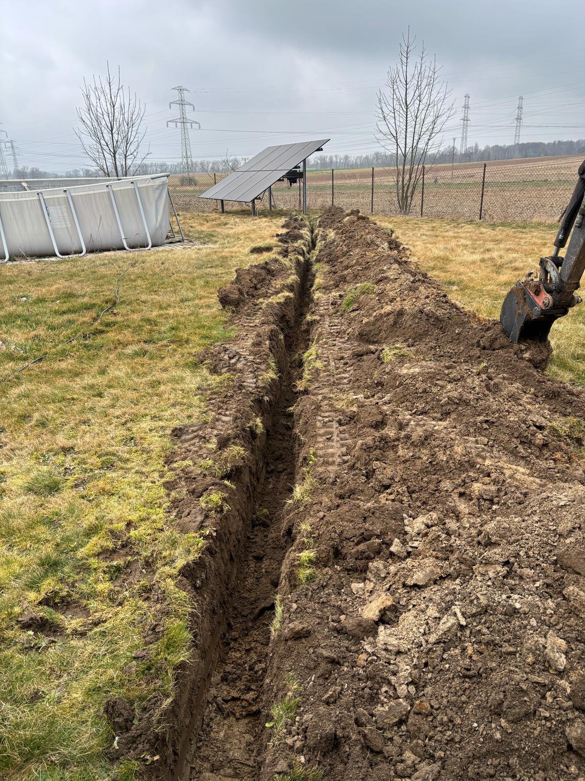 Wąski wykop w ziemi na działce z widocznym basenem stelażowym i panelami słonecznymi w tle, wykonany małą koparką.