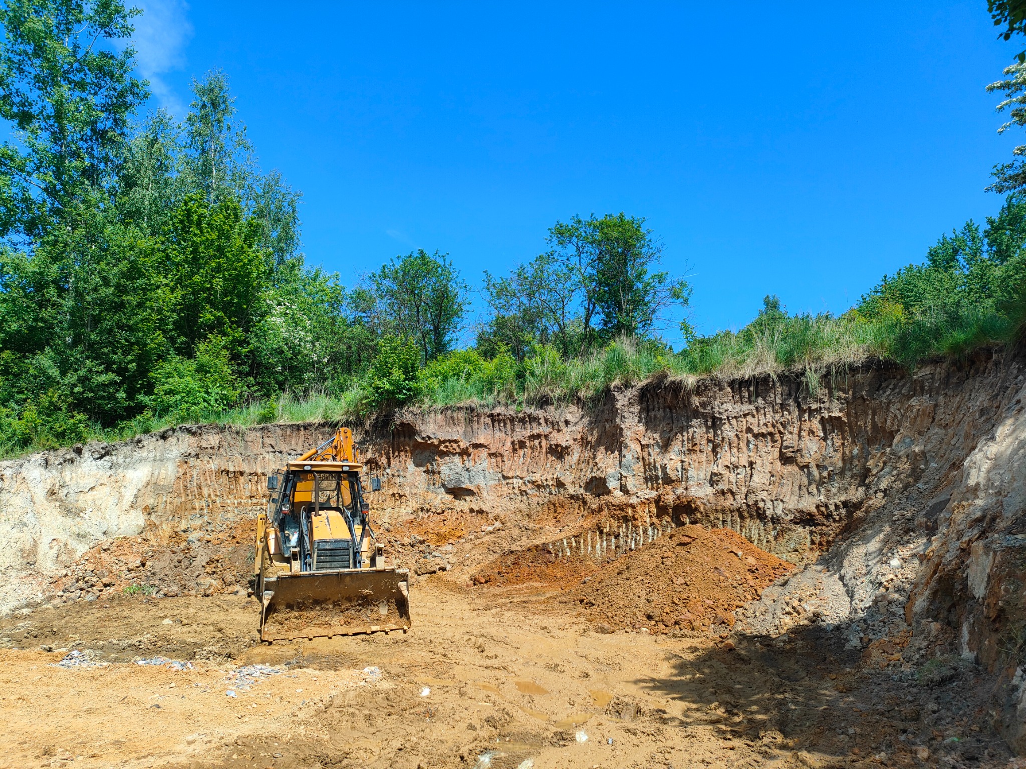 Żółta spycharka w Bogatyni pracuje na placu budowy, przygotowując teren pod inwestycję. Widoczny wykop w ziemi i drzewa w tle.