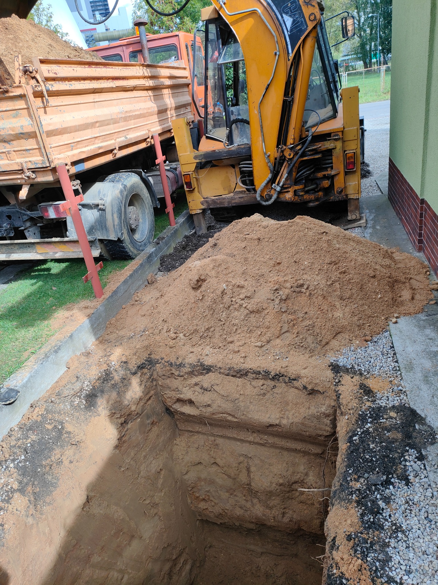 Żółta koparko-ładowarka i wywrotka obok wykopu w ziemi, widoczny fragment betonowego obrzeża i zielona ściana budynku w tle.