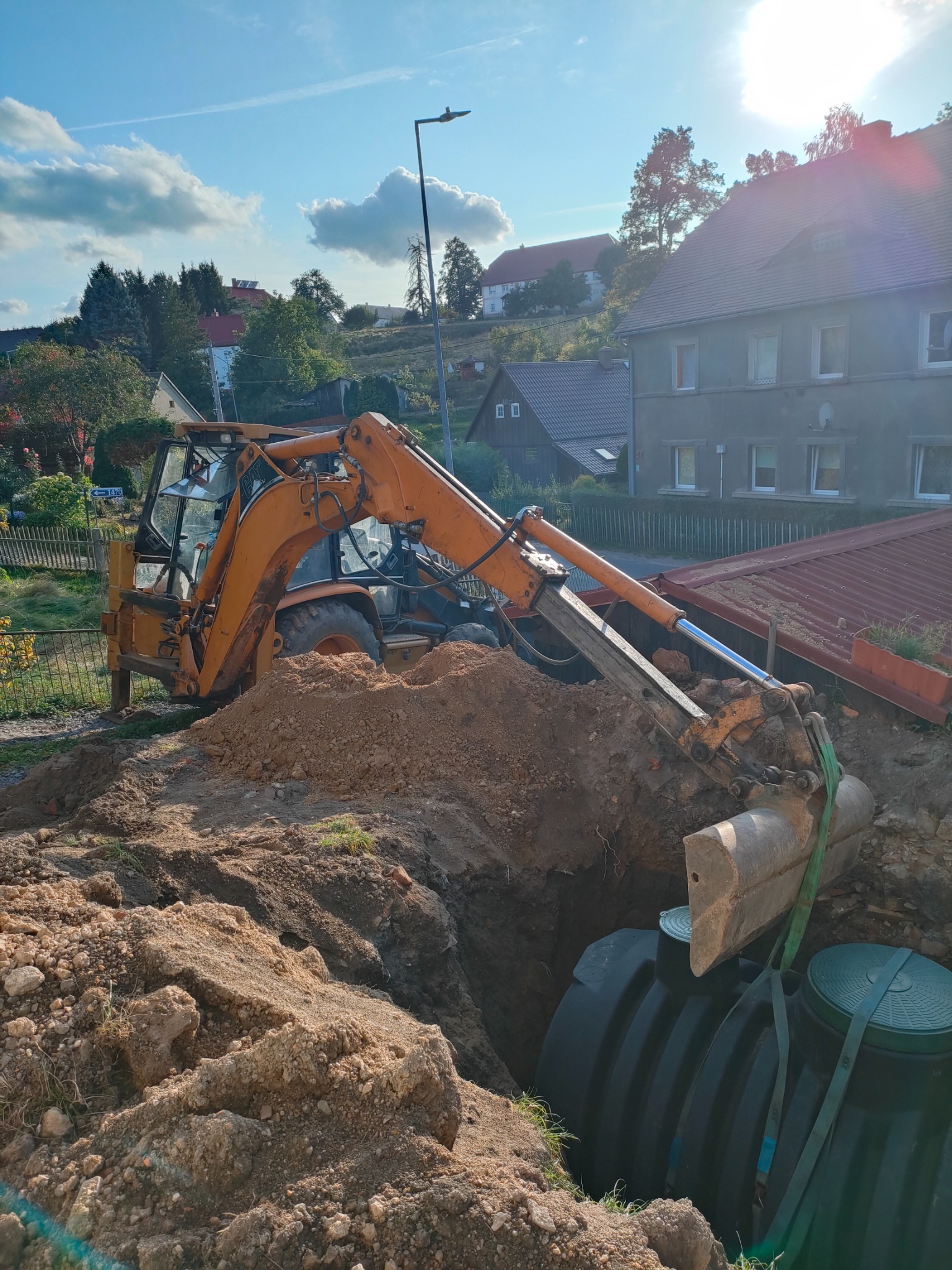 Pomarańczowa koparka umieszcza czarny, plastikowy zbiornik w wykopanym dole na tle zabudowań mieszkalnych i błękitnego nieba.