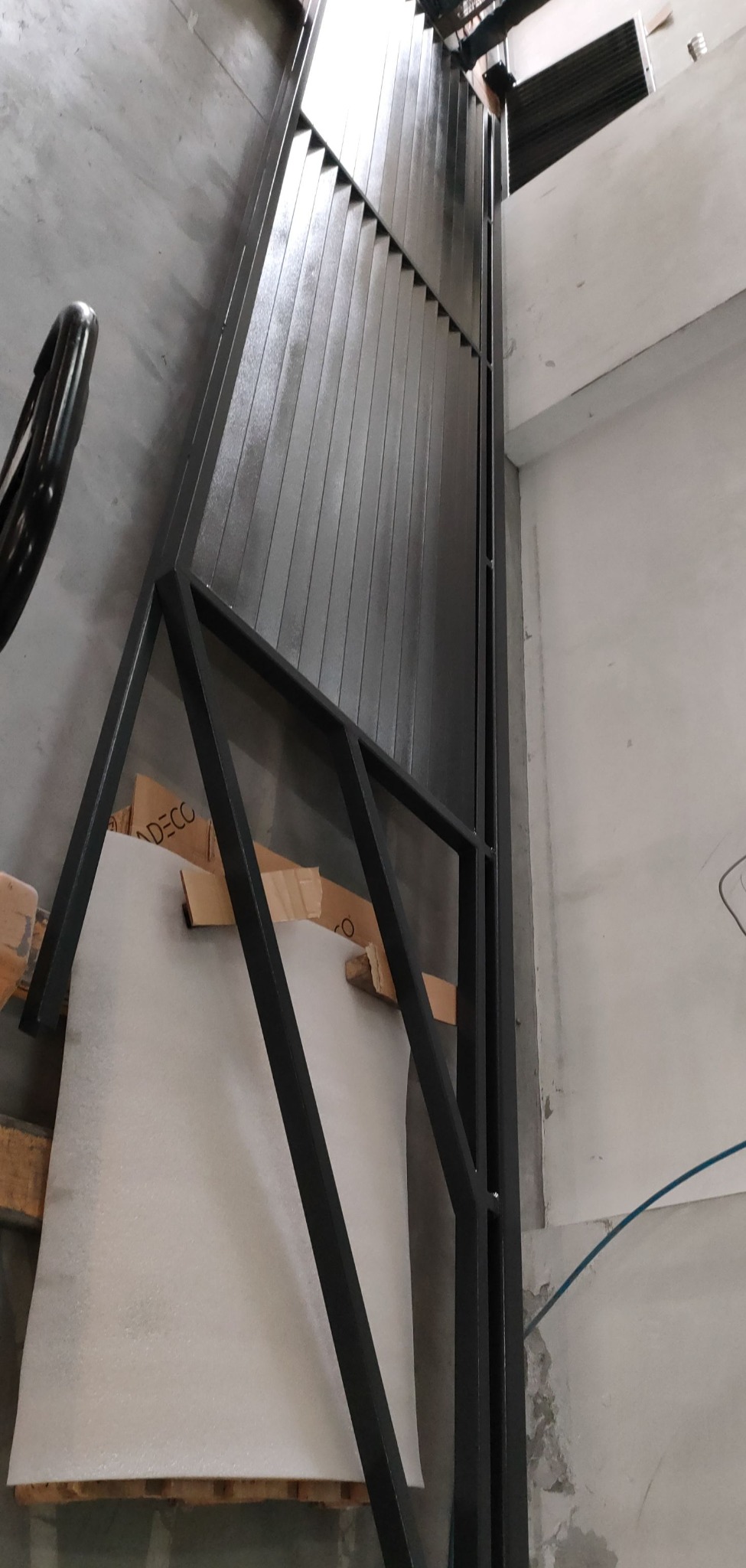Nowoczesne, czarne ogrodzenie lamelowe, częściowo zasłonięte folią ochronną, opierające się o paletę w surowym, betonowym wnętrzu.