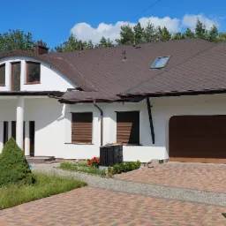 Biały dom jednorodzinny z brązowym dachem z gontów bitumicznych i brązową bramą garażową, widok z poziomu gruntu na tle zieleni i błękitnego nieba z chmurami.