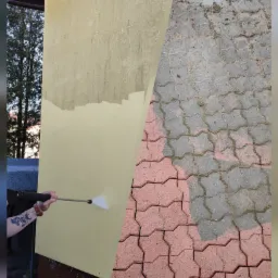 Fragment elewacji i kostki brukowej, gdzie widać różnicę w czystości przed i po użyciu myjki ciśnieniowej; na elewacji widoczne zacieki, a na kostce mech i brud.