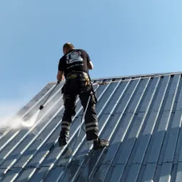 Pracownik w uprzęży, z perspektywy od dołu, spryskuje wodą pod ciśnieniem blaszany dach, widoczne krople i strumień wody, na koszulce logo firmy.