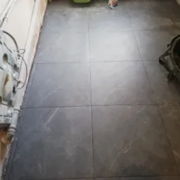 Ułożona, ciemnoszara podłoga z płytek imitujących marmur w trakcie remontu. Widoczne rury, grzejnik, wiadro i odkurzacz.