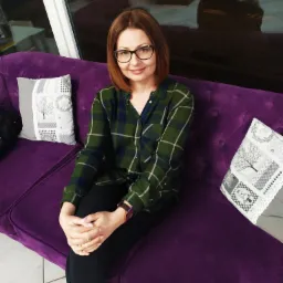 Kobieta w okularach siedzi na fioletowej sofie z dekoracyjnymi poduszkami, ubrana w zieloną koszulę w kratę i czarne spodnie, z rękami splecionymi na kolanach.