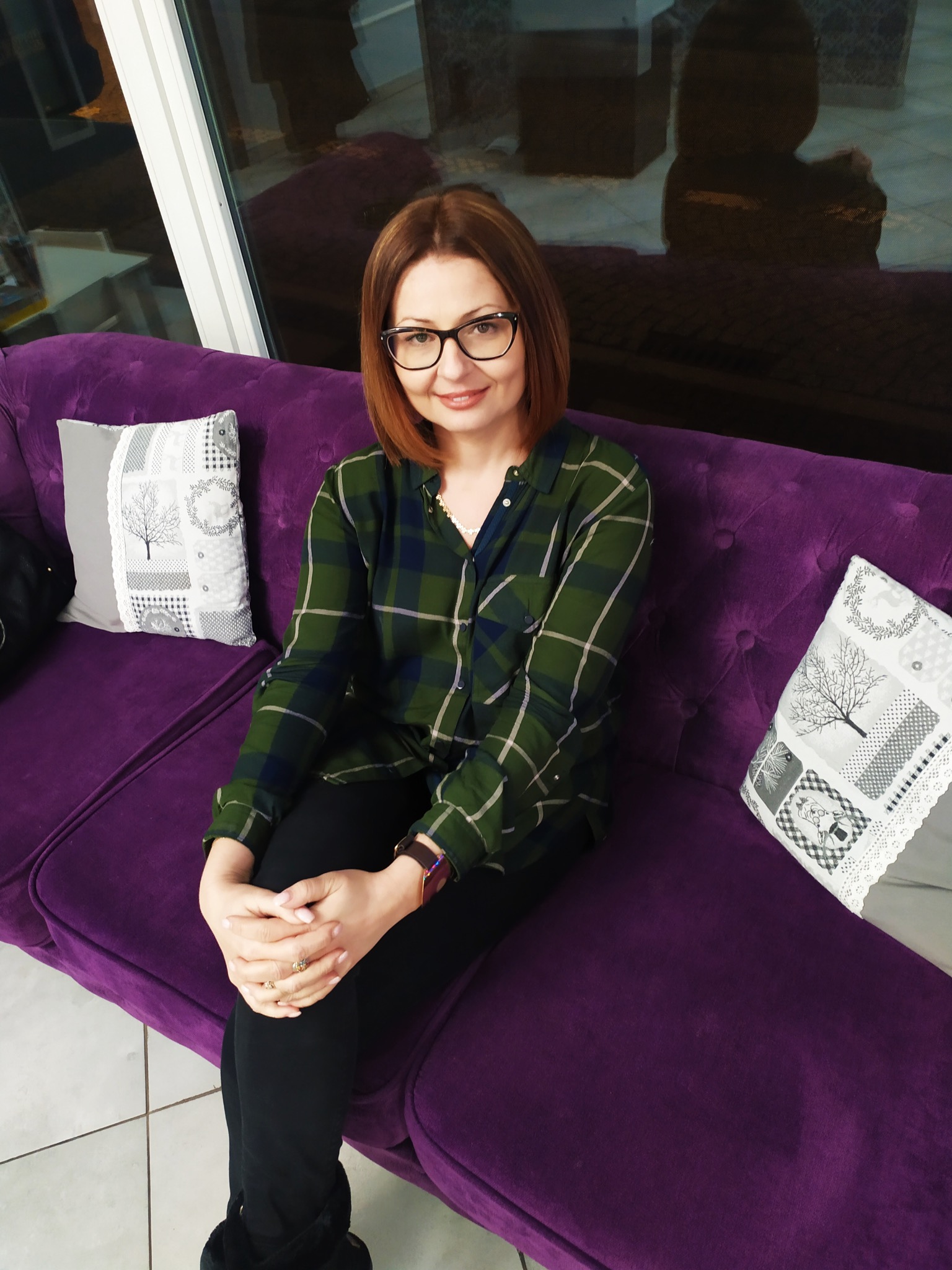 Kobieta w okularach siedzi na fioletowej sofie z dekoracyjnymi poduszkami, ubrana w zieloną koszulę w kratę i czarne spodnie, z rękami splecionymi na kolanach.