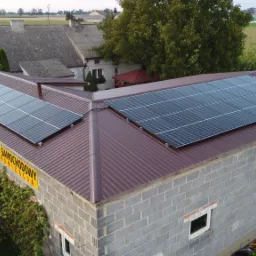 Panele słoneczne na dachu budynku warsztatu samochodowego z widocznym szyldem, otoczone zielenią i polami uprawnymi.