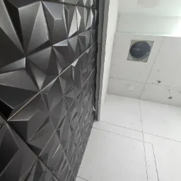 Ściana wyłożona czarnymi, trójwymiarowymi płytkami ceramicznymi w łazience, kontrastująca z białymi płytkami podłogowymi i sufitowymi, widoczny otwór wentylacyjny w suficie.